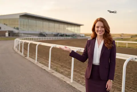 Erin Calder, the guide's author, stands at the rail of a racetrack and shows the way with an open hand, a plane climbing in the evening sky. The picture is AI-generated: she is a character, and the track is not Century Mile.